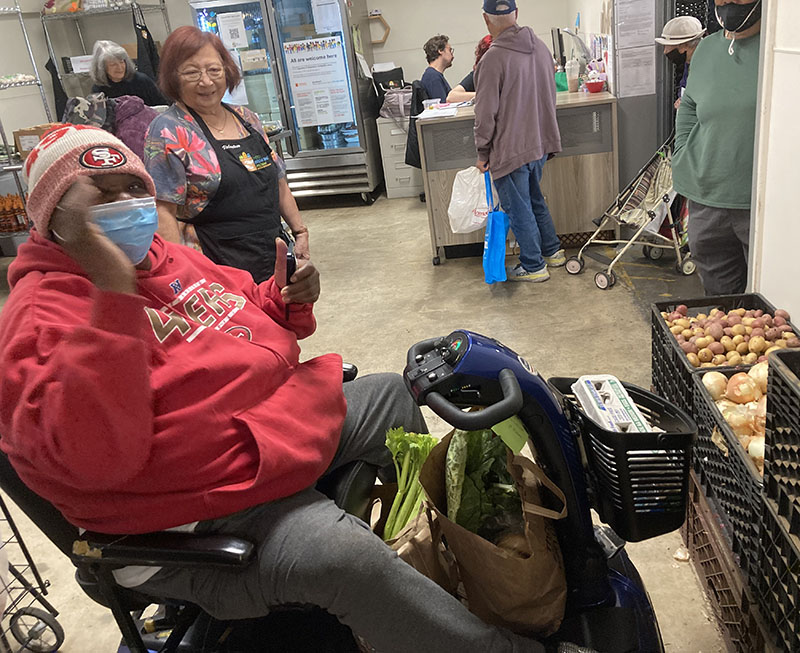 Senior volunteer helps customer shop for vegetables; other seniors check out at register.